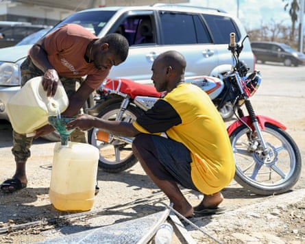A car driver and a motorcyclist transfer petrol between two jerrycans at the roadside.