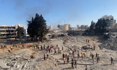 Dozens of people walking across rubble, with damaged buildings and smoke rising in the background