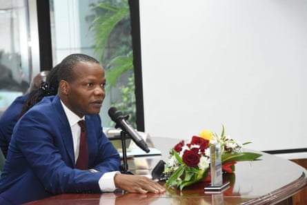 A young African man in a suit sitting at desk before a microphone