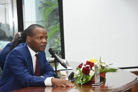 A young African man in a suit sitting at desk before a microphone