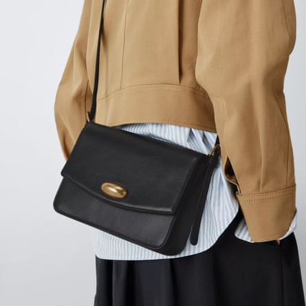 A John Lewis black crossbody bag is worn to the back by the model. The bag has a gold clasp and the model wears a long blue and white pinstripe shirt with camel coloured short jacket