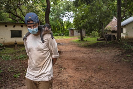 A monkey wraps itself around a man's shoulders. They are on a path through trees and small buildings