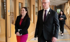 John Swinney walks with Kate Forbes down a corridor at the Scottish parliament in Holyrood; pale wood doorways and window frames are seen to either side of them.  He wears a black suit, white shirt and tie, and she wears a black jacket over a skirt which is half bright pink and half red. They both look cheerful.