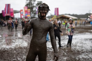 A festival-goer is wearing a full-length wetsuit and has a mask and snorkel on their head and is covered in mud. They're standing in a muddy field with other revellers and festival signs behind them