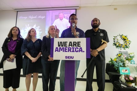 man speaks into microphone at lectern marked 'we are America' as four people stand behind him