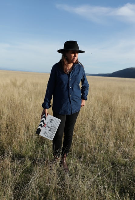 Rachel Perkins walks through grassland under a blue sky. She is wearing a broad-brimmed hat and a blue shirt and is holding a film slate in one hand