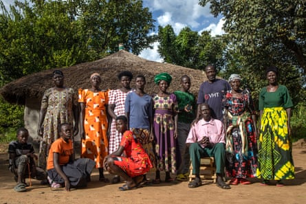 Thirteen people stand or crouch in two lines in front of a thatched building