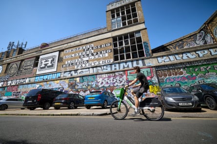 A man cycles on a Lime bike past the brick former brewery buildings, which are covered with graffiti.