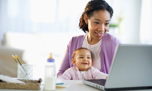 A woman holds a baby while using a laptop.