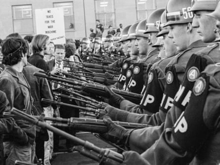 Protesters stand in front of a line of military police with their guns out