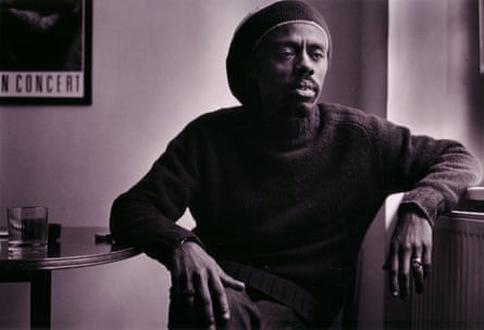 A black and white image of a man with a polo neck jumper and hat sitting in a room with his arms on a table and radiator