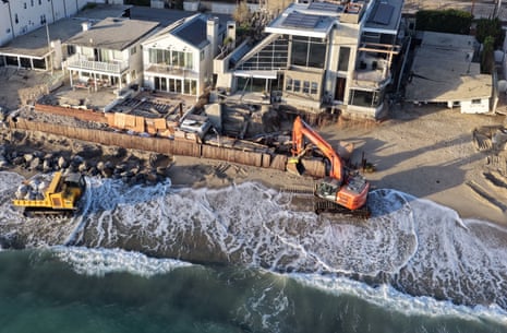 In an aerial view, a Pacific Ocean wave washes up near workers using heavy equipment to protect damaged beachfront homes with boulders.