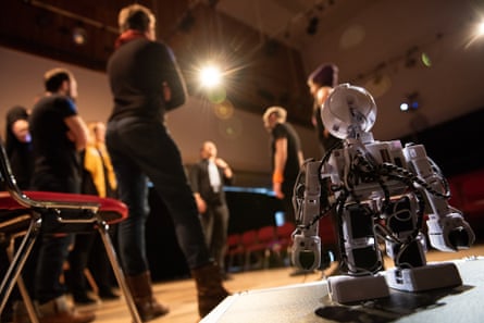 A toy robot in the foreground with an audience standing behind