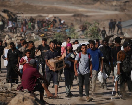 Palestinians carry aid packages: men and boys walk down a dry, dusty path in a crowd; some are carrying sacks and one has a cardboard box with a GHF logo on it