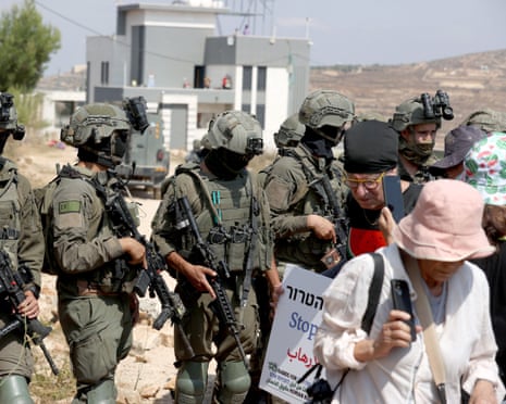 Israeli forces holding back a group of people in front of a house with people on the terrace