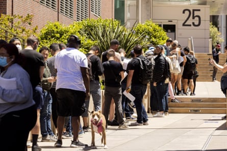 People stand in a long line leading up the steps to a building. One man has a dog on a leash with him.