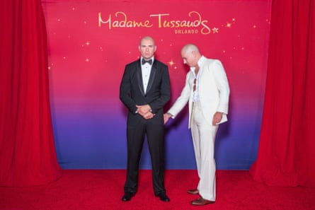 Pitbull, in a white suit, touches a wax figure of himself wearing a black suit. The background, which starts off red and fades to purple, reads ‘Madame Tussaud’s’ and is flanked by red curtains.