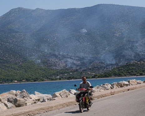A local on motor bike with plumes of smoke in the hills behind them as firefighting teams try to stamp out pockets of fire in Psatha, Greece last week.