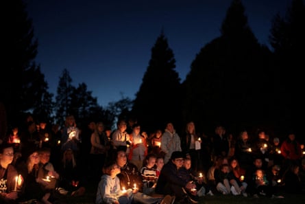 Mourners hold candles at a vigil for shooting victims, trees silhouetted at dusk