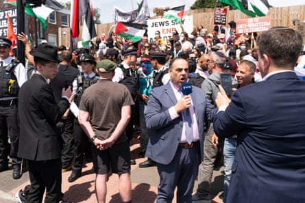 A reporter holding a microphone delivers a report with a group of protesters, some of them waving Palestinian flags, behind him.