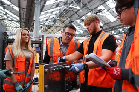 Andy Burnham in an orange hi vis with three students showing him their practical work at the factory.