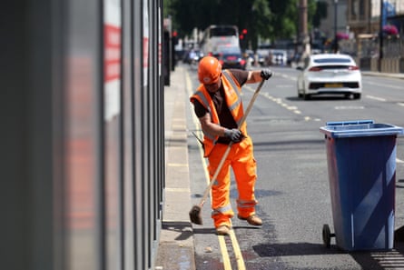 A man wearing hi-vis and a construction helmet sweeps the street outside a construction site in London.