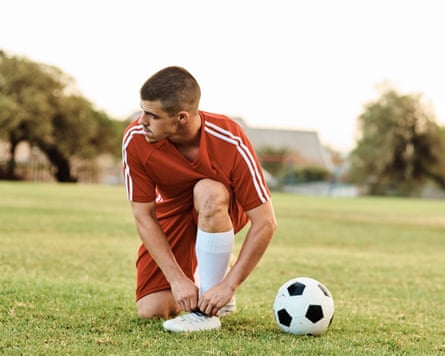 A man in football kit on a grass pitch kneels by the ball to tie the laces of his boots
