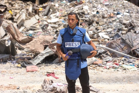 Anas al-Sharif, wearing a blue press protection vest and holding a microphone, standing next to the rubble of a bombed building