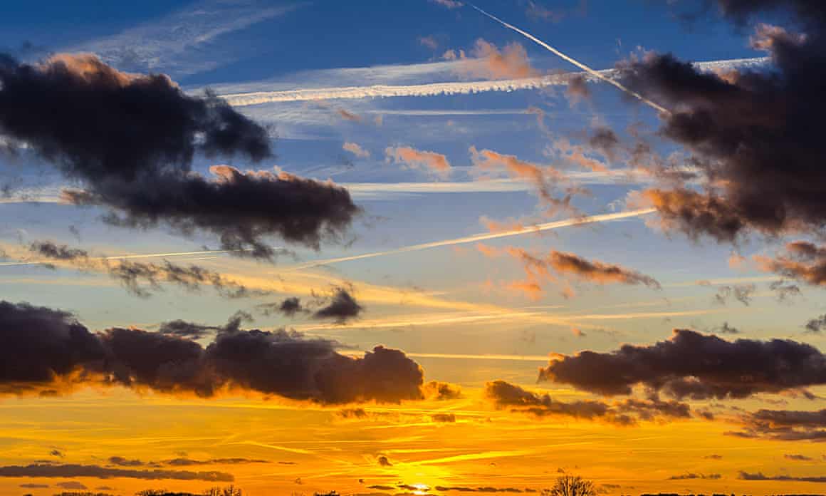 The sky streaked with aircraft contrails is photographed at sunset, its fluffy clouds dark in the light.