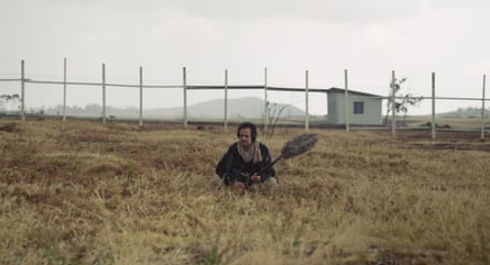 A man holding a boom microphone sits in a field in a rural landscape.