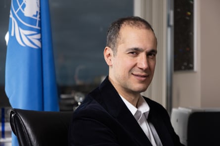 A youngish man in a suit jacket smiles at the camera with the UN flag behind him