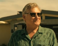 A man in a green shirt wearing sunglasses with a cabin in the background