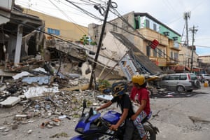 Two people on blue motorbike look at debris from a collapsed building, part of which has crushed the front of a silver SUV