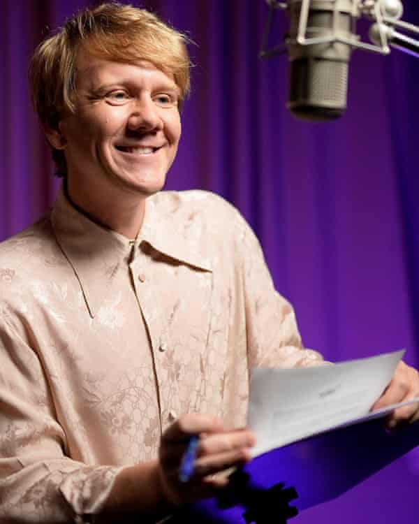 Josh Thomas in a promo shot for his 2022 Audible podcast How To Be Gay, he is smiling and wearing a pale pink shirt, holding a piece of paper in front of a microphone