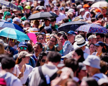 A large crowd of people some holding up umbrellas to shade themselves from the sun