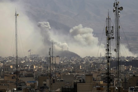 Wide shot with smoke in distance over urban scene with what appear to be communications pylons in foreground