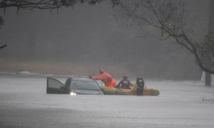 A yellow SES swift water rescue boat with two volunteers in it attends to Brian Russell’s car which is partially submerged in flood waters with one door open. An SES member wearing bright orange is leaning out of the boat towards the car while the other member drives the boat