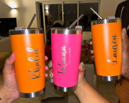 Three hands holding sippy cups – orange, pink, orange – with metal straws in, with names in cursive writing on the side of each one