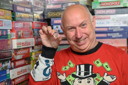 A man wearing a Monopoly jumper holds the coin, in front of rows of Monopoly board game editions.