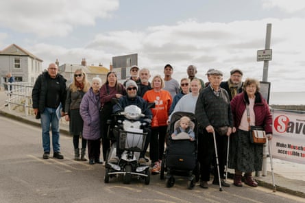 A group of about 12 residents at a bus stop in Cornwall, one on a mobility scooter, some with walking sticks, on the side of the street with a ‘Save our Bus Stop’ sign
