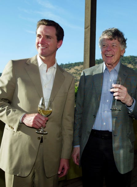 Gavin Newsom and Gordon Getty standing outside, side by side, both wearing jackets and shirts with no ties, and holding glasses of wine, laughing and look to the left of the camera, against a blue sky with a hill in the background