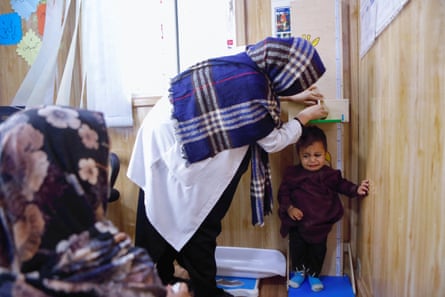 A woman in a headscarf and white lab coat measures a crying child as another woman looks on