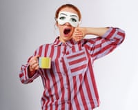 Woman in colourful stripey pyjamas yawning, holding a mug and wearing an eye mask with eyes painted on it