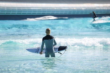 Vaughan stands in the shallows of the wave pool holding his board and looking out to a surfer on a wave