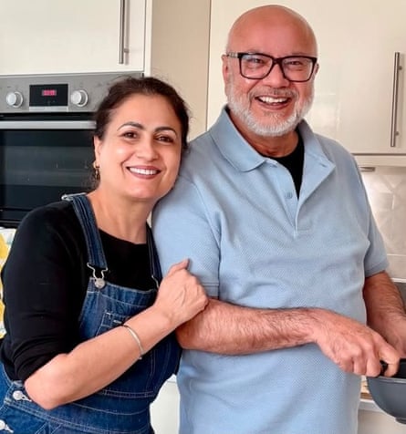 Avtar Singh wearing a light-blue polo shirt, cooking at a hob, while Nicci Dhamu leans her head on his shoulder with her left arm around his back and her right hand on his right arm, both smiling at the camera