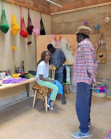 A woman sits on a chair underneath string bags full of coloured string, next to a table holding artists’ tools and materials, while a man looks on