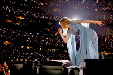 A woman in a powder blue dress leans over an audience of many people spotlit, singing into a mic