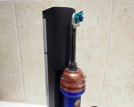 A toothbrush-style attachment sits on a Dyson vacuum cleaner docked against a tiled wall