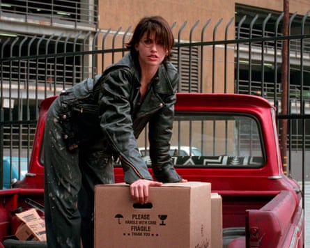 Wearing a leather jacket and workwear trousers, she is standing on the back of a pickup truck, leaning down to pick up a large cardboard box