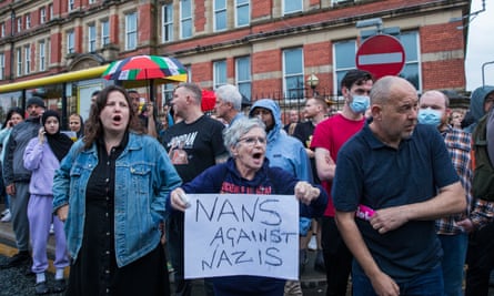 People stand together on a road in front of a large red-brick building shouting, with one woman holding a sign saying ‘Nans against Nazis’
